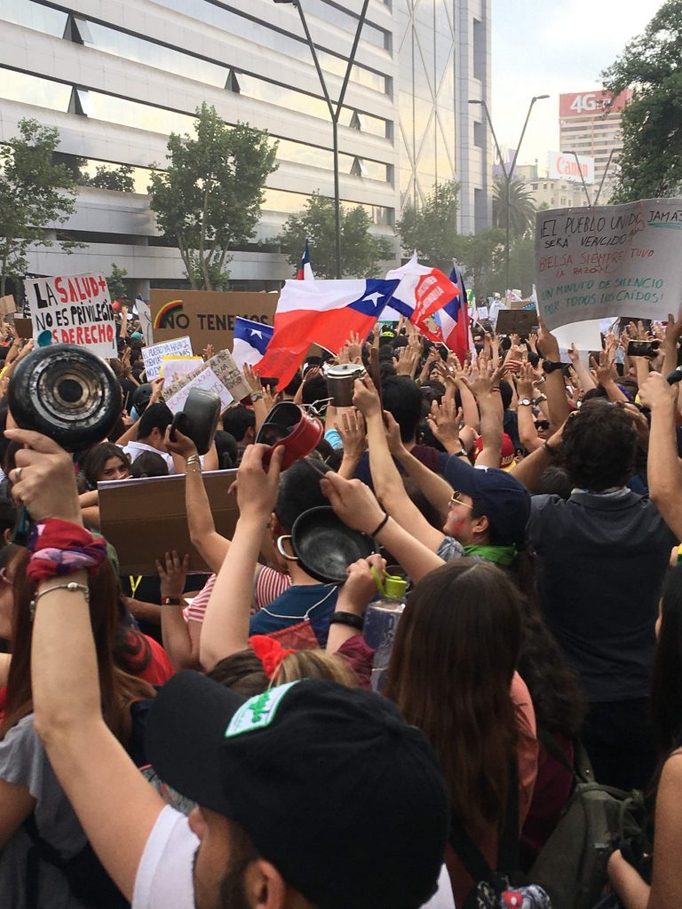 Manifestantes caceroleando, al fondo varios carteles y banderas chilenas. Marcha 25 de octubre de 2019 por Av. Providencia.