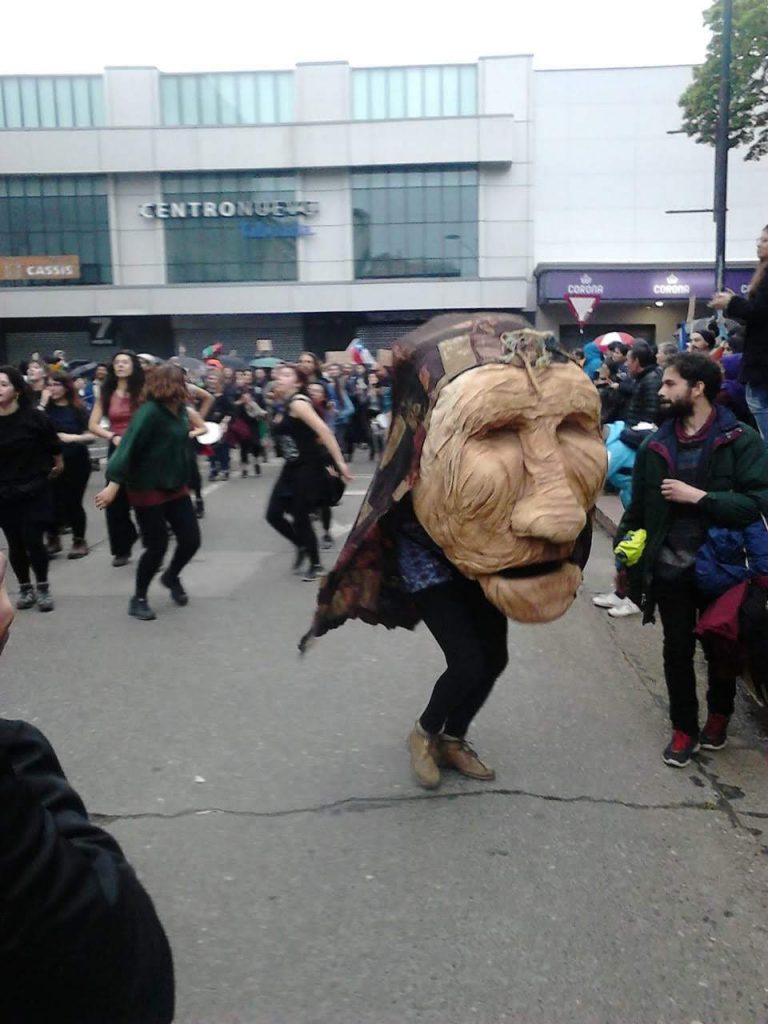 Manifestación en Plaza de Armas de Valdivia. En primer plano, alguien baila con una cabeza gigante con rasgos indígenas.